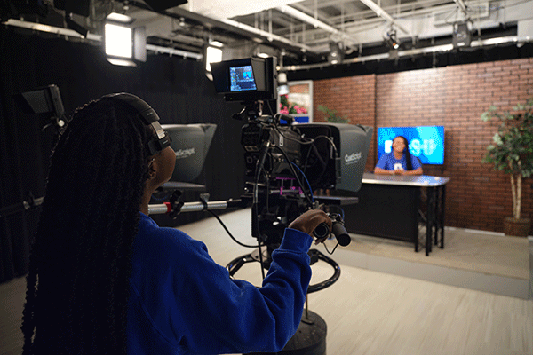 Inside a media newsroom, a Black student with long black dreadlocks stands behind a black camera and teleprompter, wearing a blue sweatshirt. On the set is a Black student wearing a blue T-shirt, blurred in the photo. A desk and television are visible on the set. A brick wall is in the background. Ceiling lights are also visible in the newsroom.