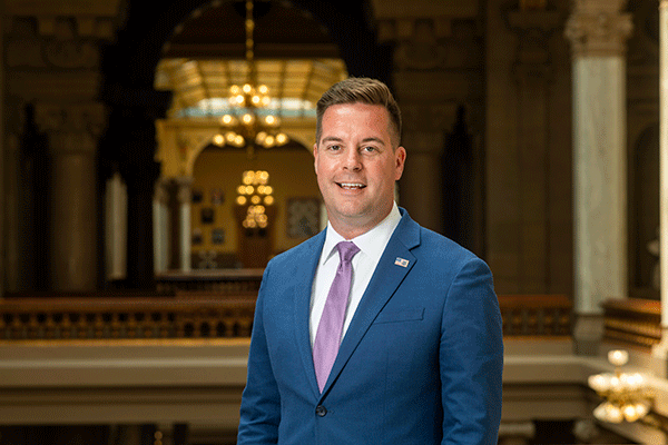 White male with brown hair dressed professionally with suit coat and standing against a railing on the second floor of the Indiana Statehouse in Indianapolis.